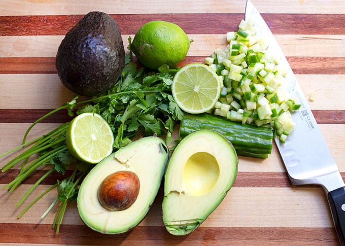 Two avocadoes, cilantro, cucumber, and limes on a cutting board