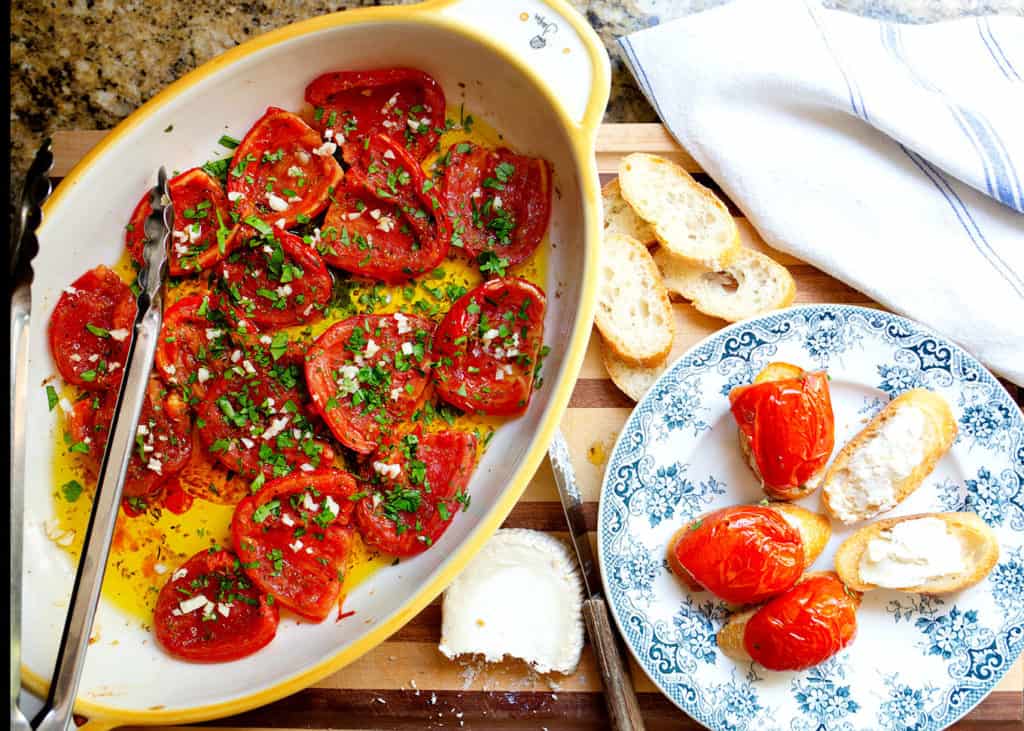 Baking dish with finished tomatoes, slices of bread with goat cheese spread on them, topped with the tomatoes.