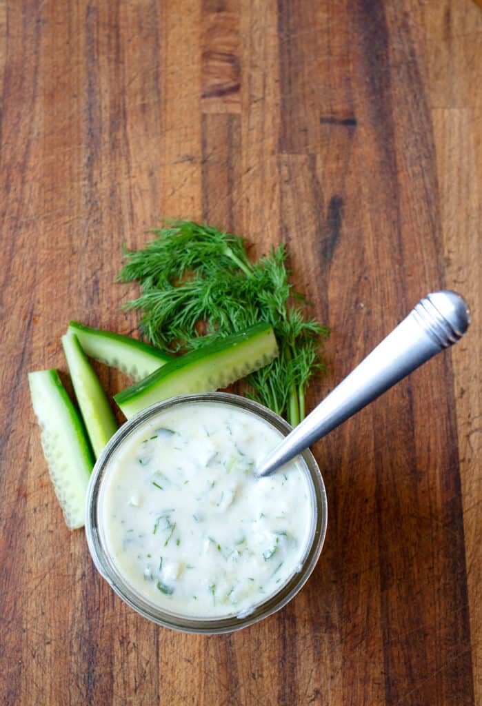 A small jar pf Tzatziki sauce with cucumber spears and fresh dill laying on the board beside