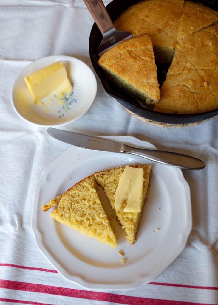 A wedge of cornbread with butter next to a skillet of cornbread and a dish of butter