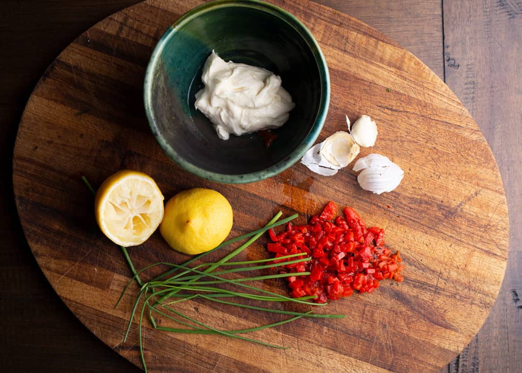 Bowl with mayonnaise on a cutting board with lemon halves, chives, roasted red peppers, and garlic cloves