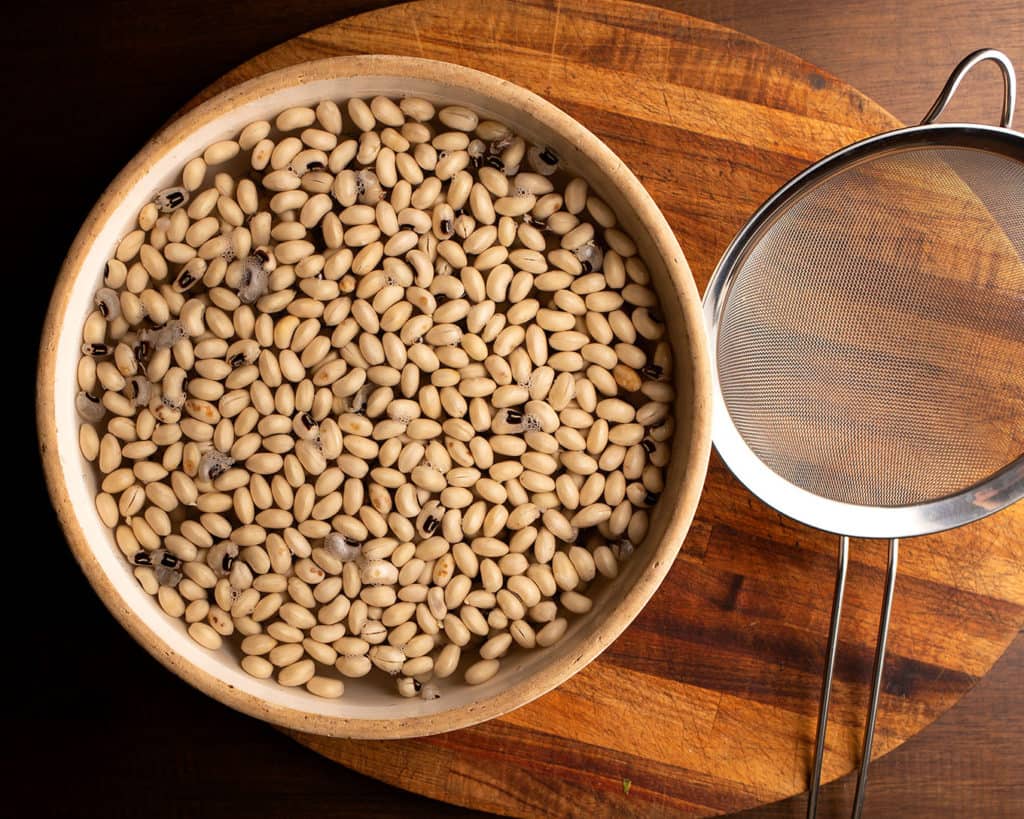 Soaked black-eyed peas in a large pottery bowl with a strainer beside