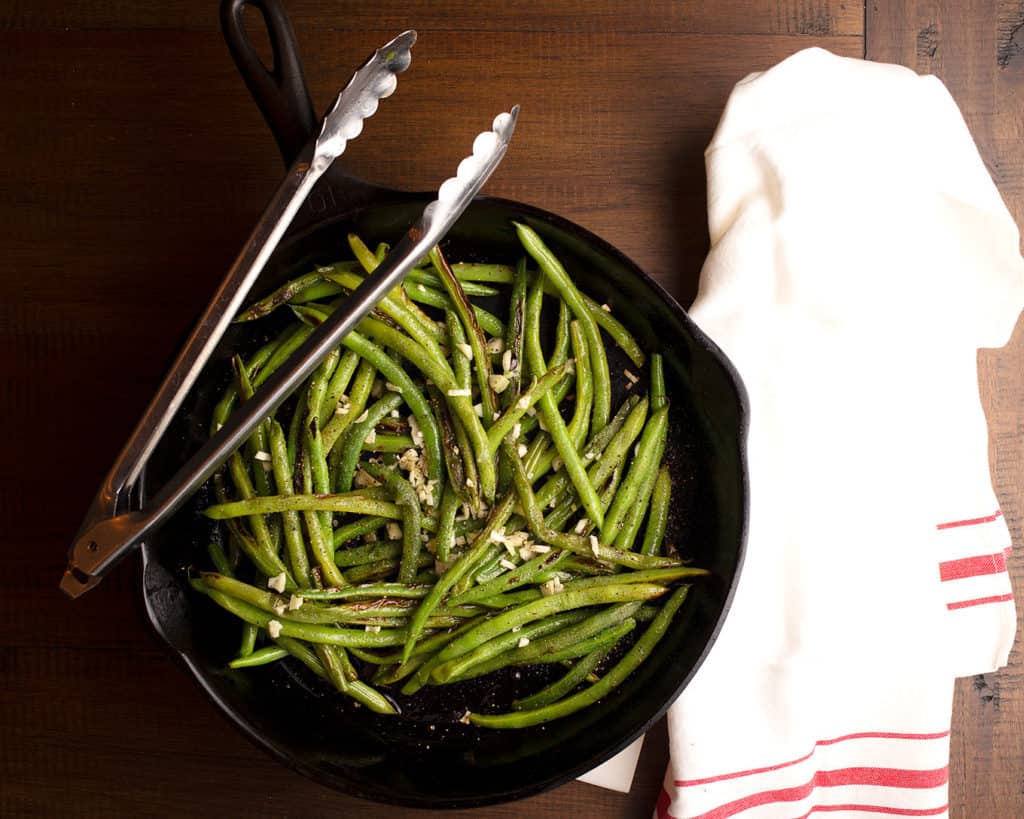 Skillet full of Garlic Green Beans with a pair of tongs and a kitchen towel
