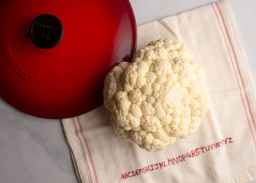 Whole head of cauliflower on a kitchen towel with a red Le Creuset lid laying beside