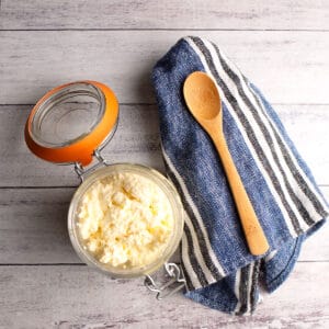 Jar of homemade ricotta cheese with a small wooden spoon beside on a blue striped napkin