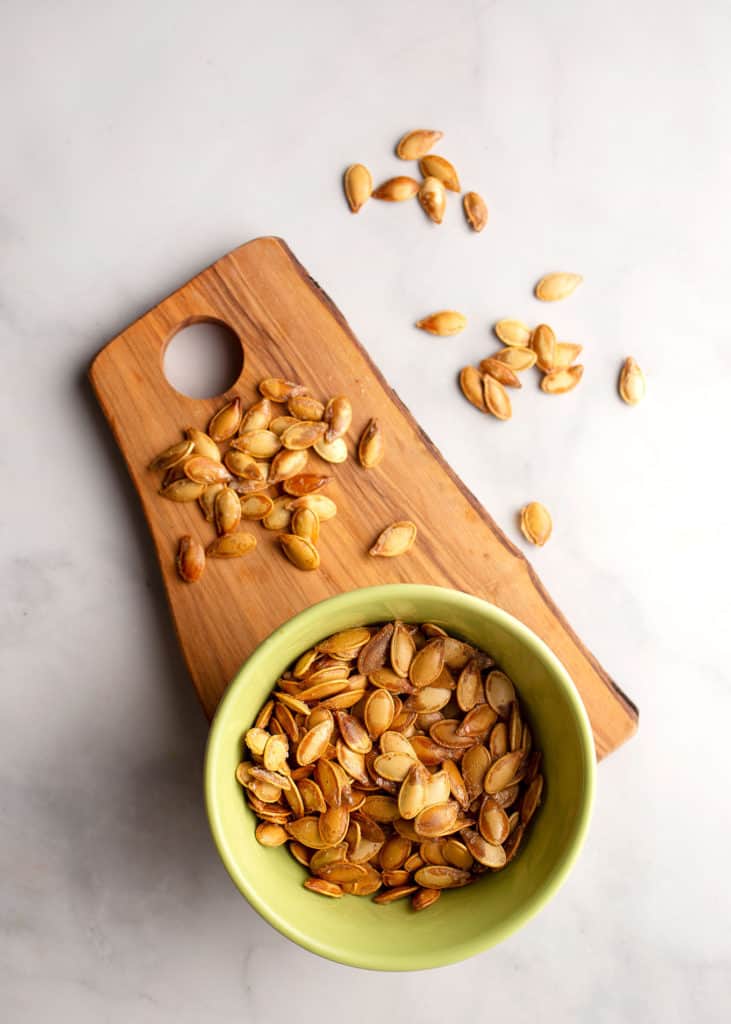 Bowl of roasted pumpkin seeds on a small cutting board with other seeds strewn about