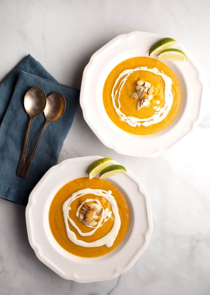 two servings of Southwest Pumpkin Soup with cream drizzled, pumpkin seeds, and lime wedges