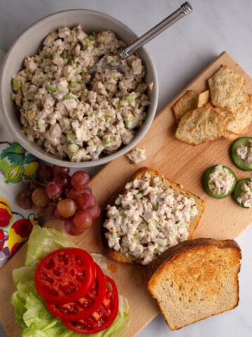 A bowl full of Honey Almond Chicken Salad with a sandwich on a cutting board, some crackers, chicken salad on cucumber rounds, lettuce and tomatoes, and a bunch of grapes.