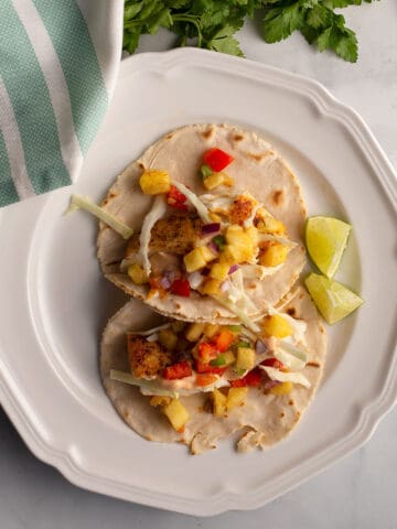 Two halibut tacos garnished with pineapple salsa on a white plate with a striped towel beside