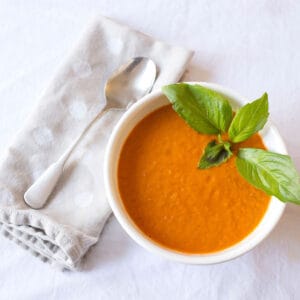 A white bowl filled with Roasted Tomato Soup garnished with basil leaves and a soup spoon beside the bowl on a napkin