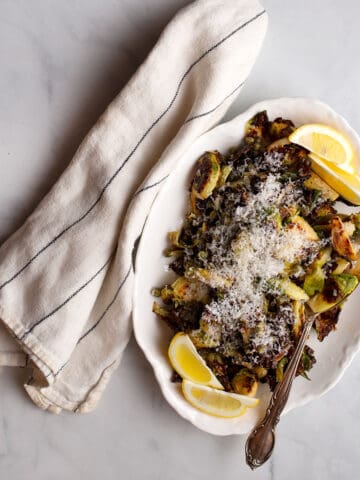 A white platter full of charred lemon caper brussels sprouts with lemon wedges for garnish