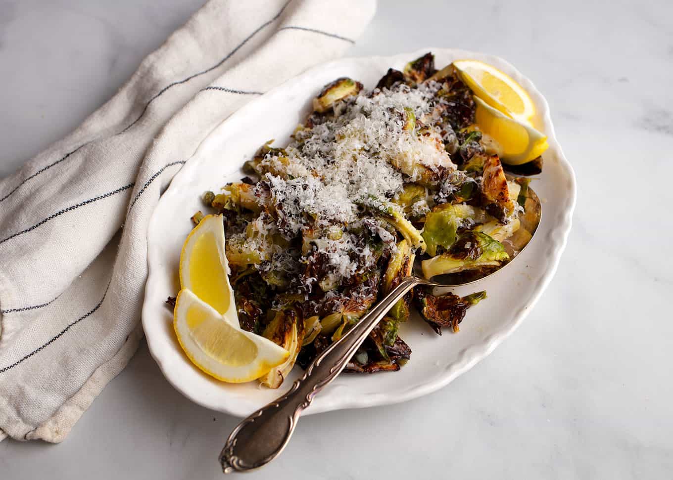 A white platter full of charred lemon caper brussels sprouts with lemon wedges for garnish with a vintage silver serving spoon