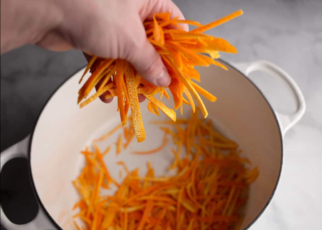 A hand dropping julienned orange peels in the saucepan