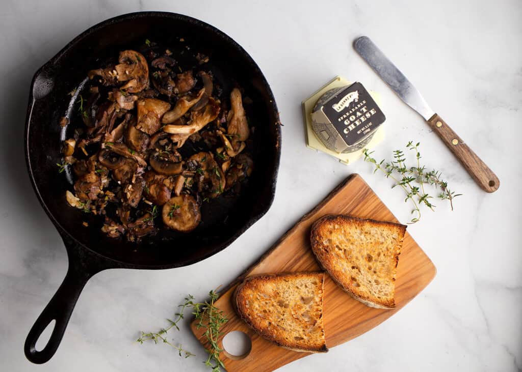 Skillet of sauteed mushrooms, board with toasts, and goat cheese container