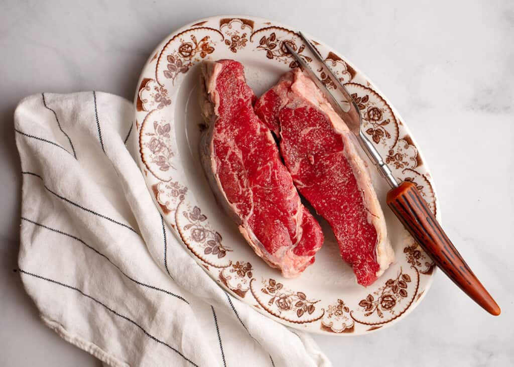 Grass-fed New York strips seasoned and coming to temperature on a vintage platter