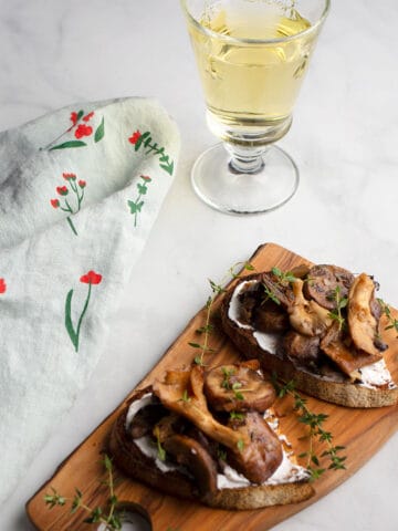 two mushroom tartines on a small cutting board with a glass of white wine