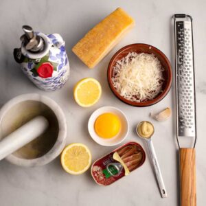 All of the ingredients for a Caesar salad dressing: freshly grated parmesan cheese, lemon, mustard, egg yolk, anchovies, garlic, olive oil