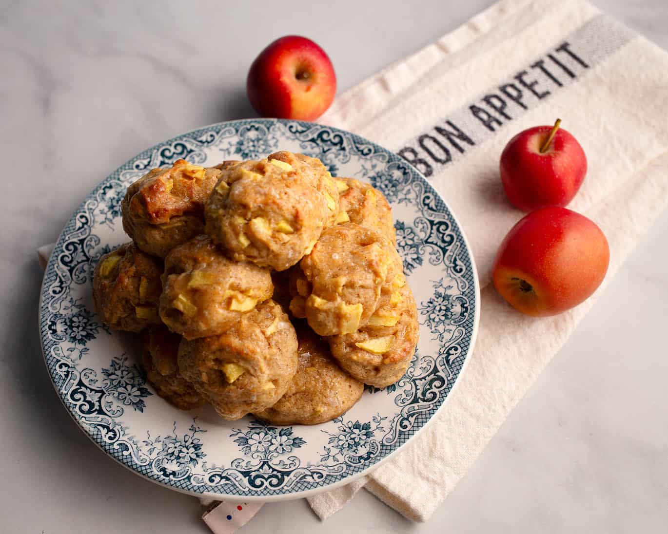 A stack of glazed Apple Fritters on a blue transferware plate with three red apples on a Bon Appetit tea towel.