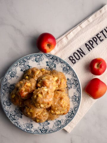 A stack of glazed Apple Fritters on a blue transferware plate with three red apples on a Bon Appetit tea towel.