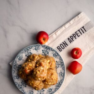 A stack of glazed Apple Fritters on a blue transferware plate with three red apples on a Bon Appetit tea towel.