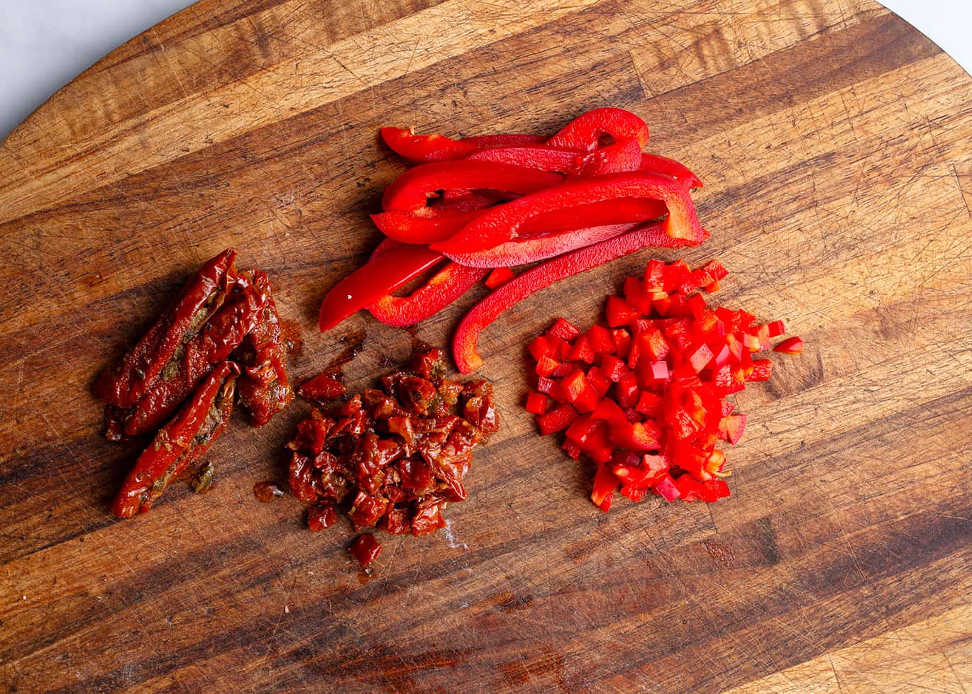 Chopped red pepper and chopped sundried tomatoes on a wooden board.