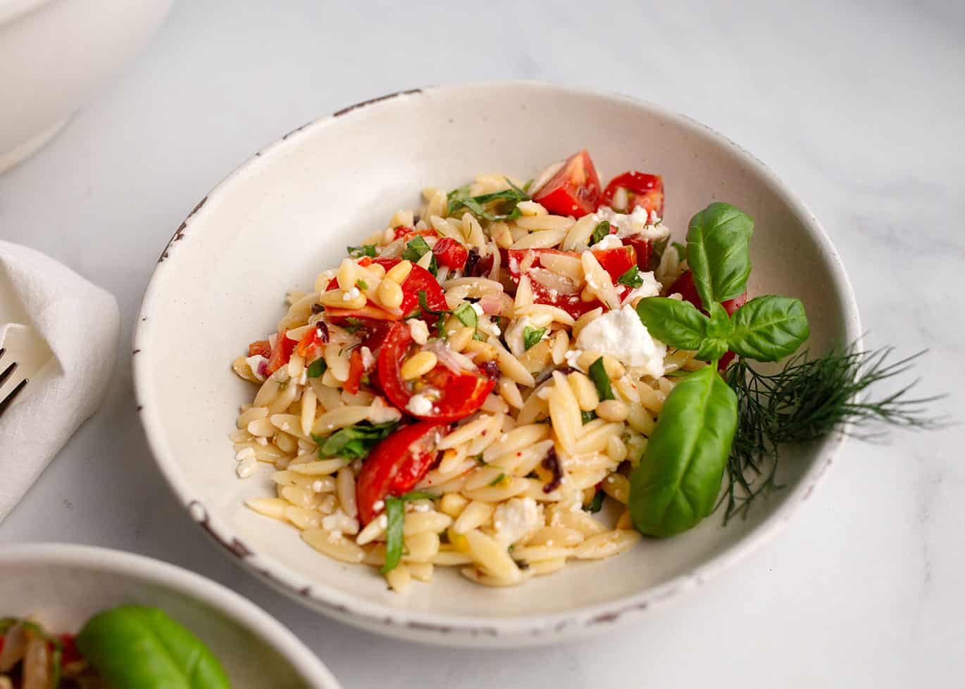 Close up of a pottery bowl of Mediterranean Orzo Salad garnished with basil and dill.