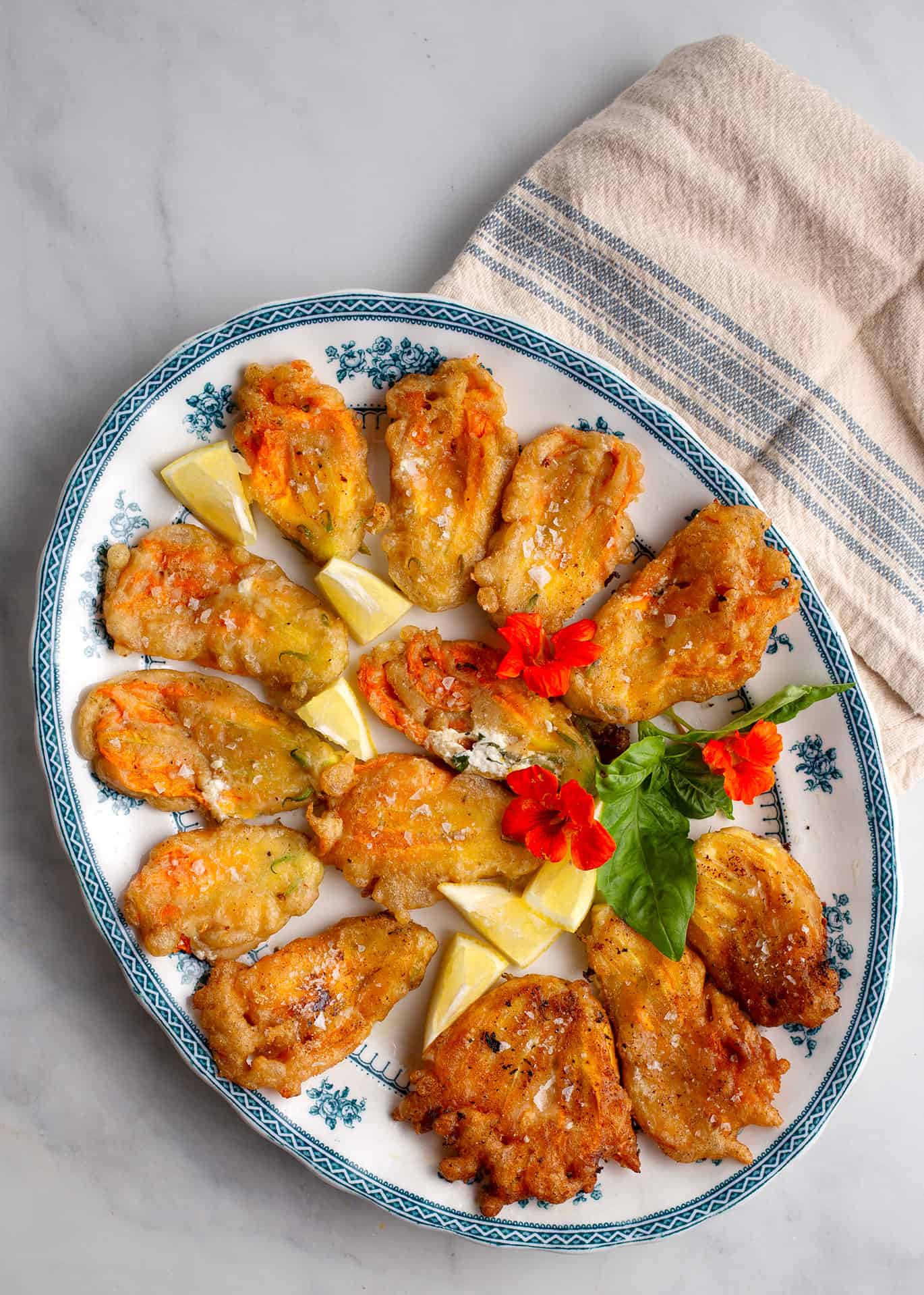 Lemon Ricotta Stuffed Zucchini Flowers on a blue and white platter with lemon wedges, basil, and edible flowers.