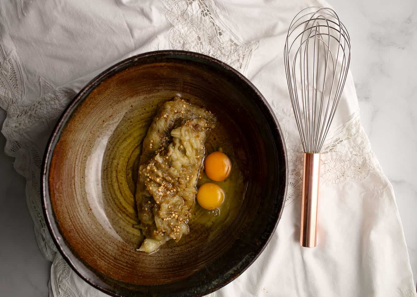 Pottery bowl with eggplant, oil, eggs, and vanilla with a copper handled whisk beside to mix with.